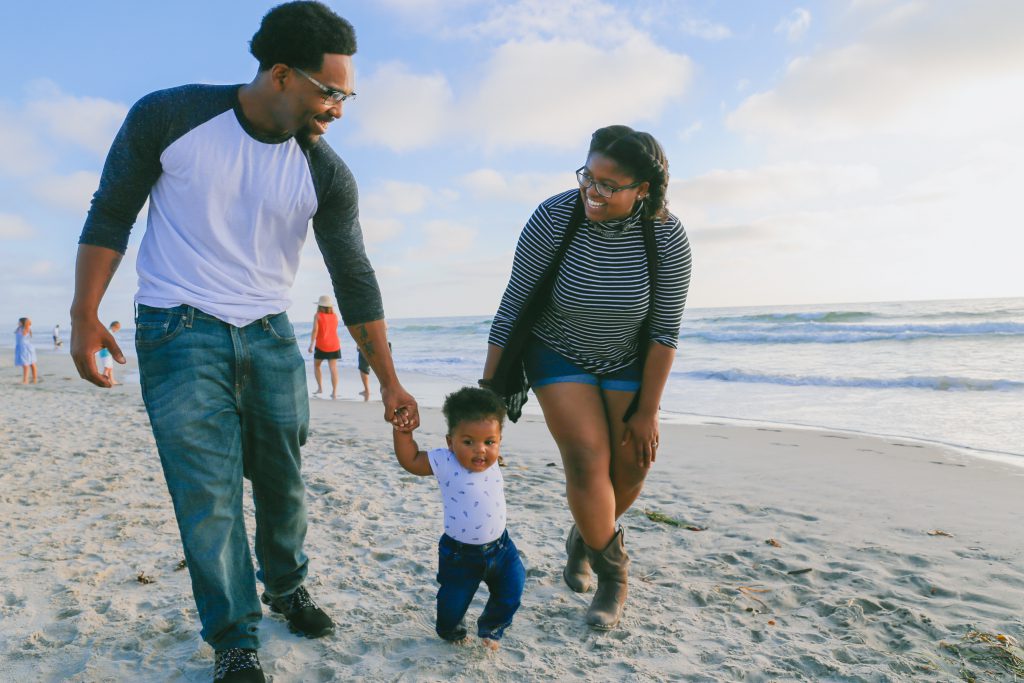 Two parents hold the hands of a toddler as they walk on the beach