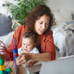 A woman looks at her computer while a child on her lap plays