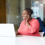 A woman sitting at a computer desk smiles as she talks on the phone