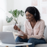 A woman wearing a headset and holding a notebook and pen talks on a video call using her laptop