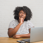 A woman looks into the distance thoughtfully while sitting at a desk with an open laptop