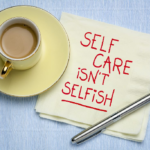 A cup of tea on a yellow saucer sits next to a napkin that says, "Self care isn't selfish"